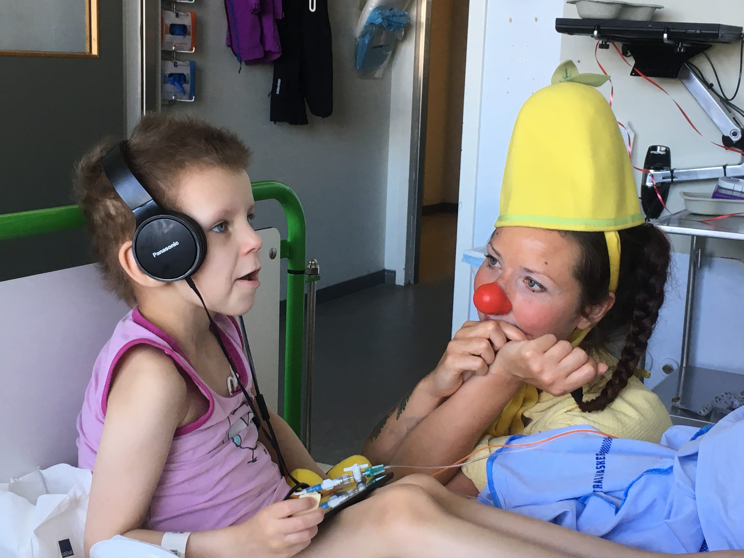 Signe sidder i en hospitalsseng med høretelefoner på, mens hospitalsklovnen Citronella sidder ved siden af