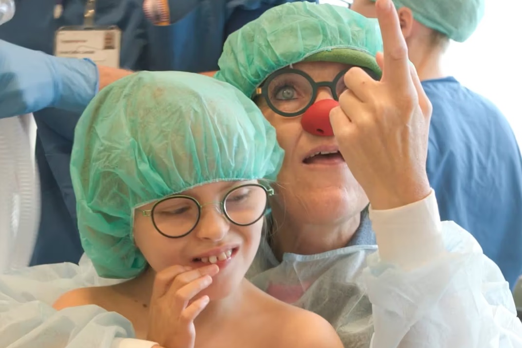 Jonathan og hospitalsklovnen Luna i operationsforberedelse, iført grønne operationshuer – Luna peger op og får Jonathan til at smile.