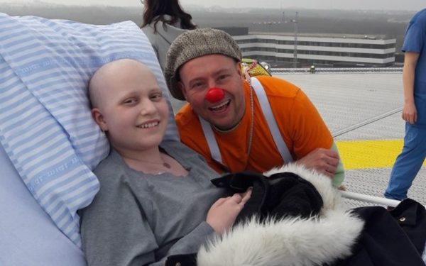 Clara ligger i en hospitalssengen og hospitalsklovnen Theodor står ved siden af. De står på toppen af en helikopterlandingsplads på et hospital.