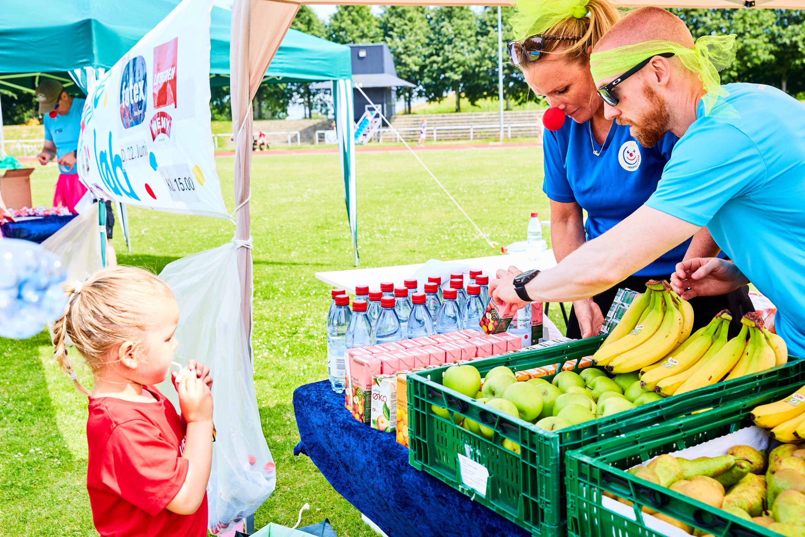 Pernille og Sidney hænger plakater op til Klovneløb. De står foran en rød mur og smiler til hinanden, mens de hænger en gul plakat op.