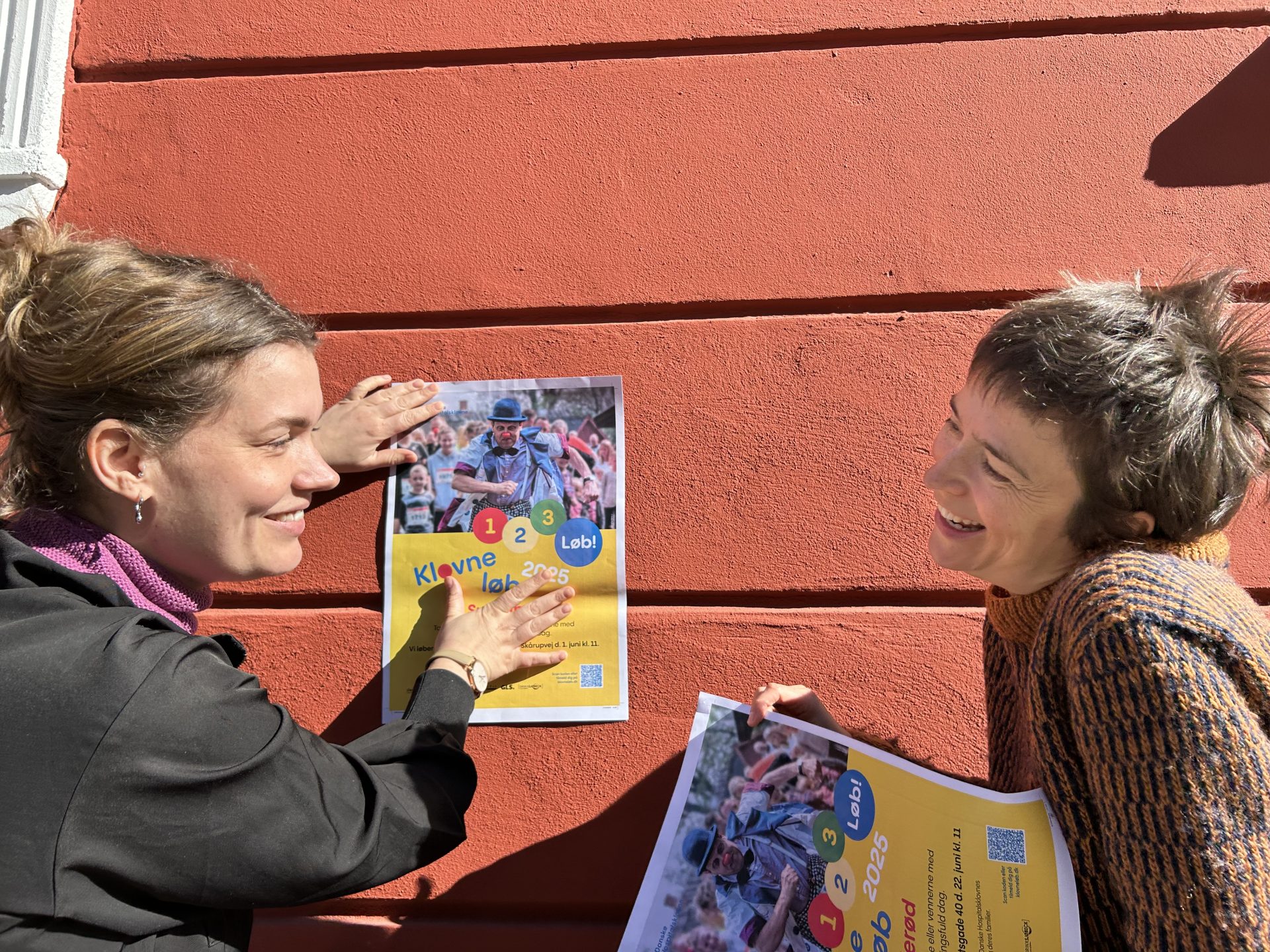 Pernille og Sidney hænger plakater op til Klovneløb. De står foran en rød mur og smiler til hinanden, mens de hænger en gul plakat op.