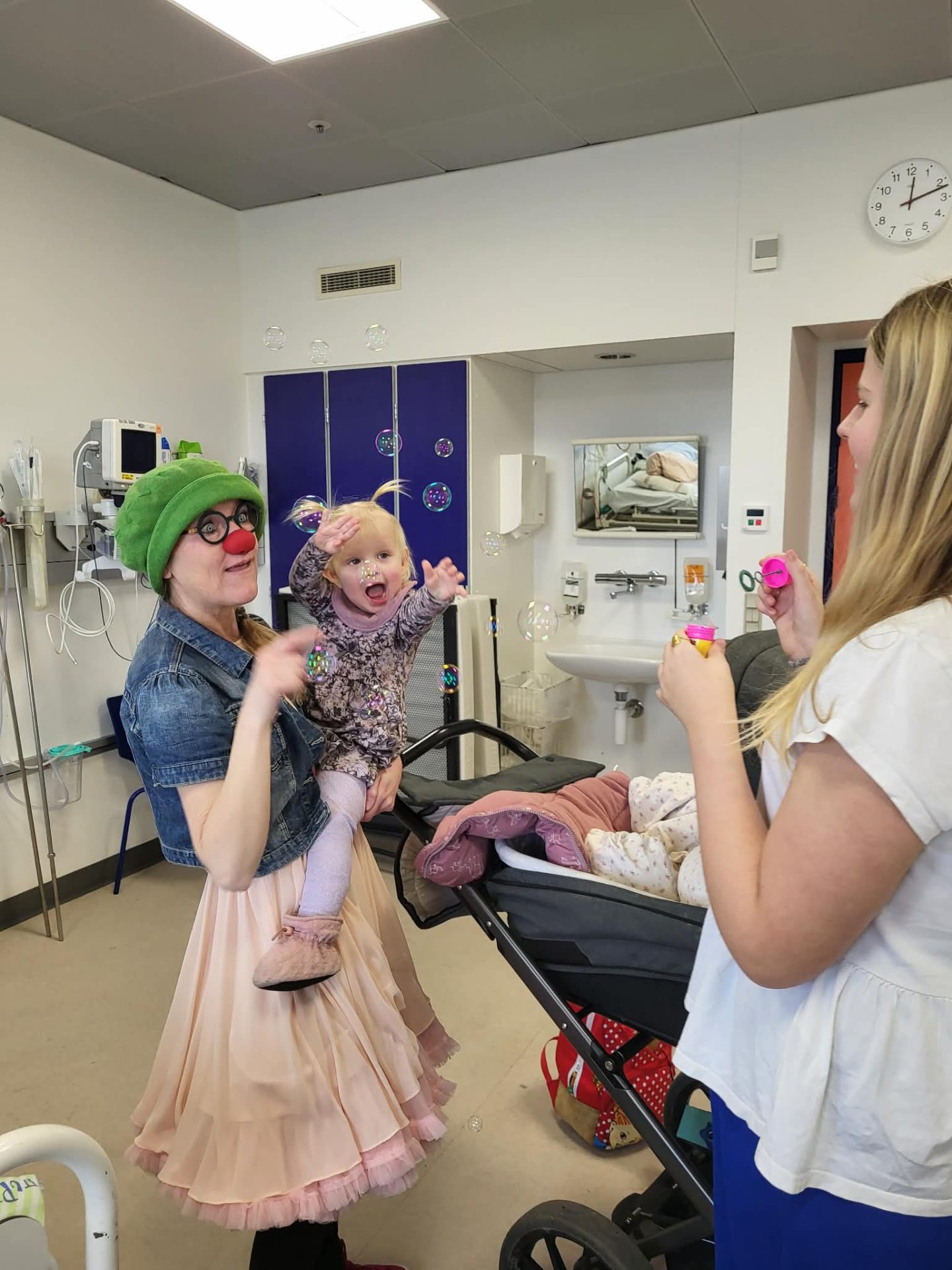 Hospitalsklovnen Luna holder Mai i armene, mens storesøster Josefine puster sæbebobler. De er på en hospitalsstue.