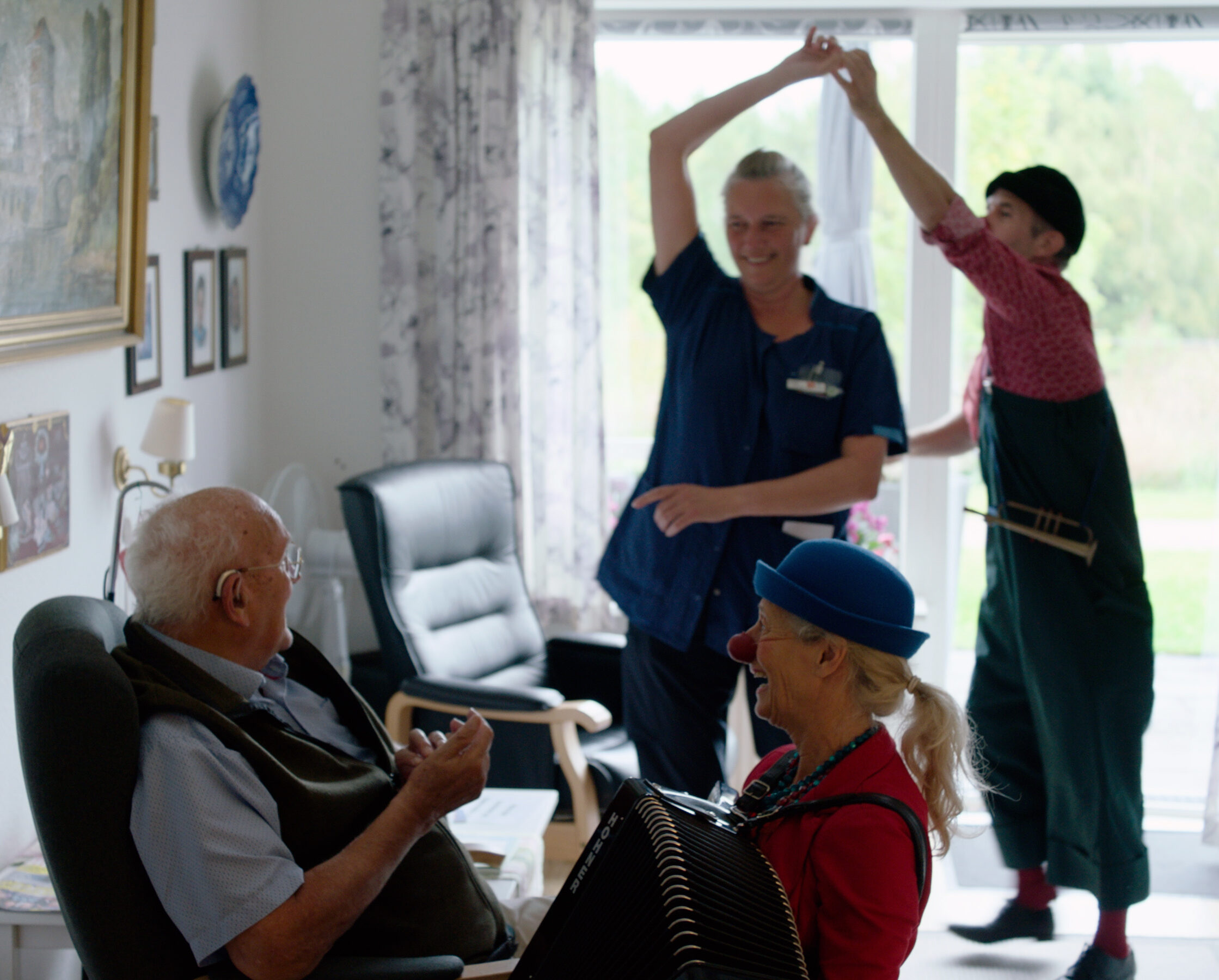 Hospitalsklovn spiller harmonika for ældre mand, mens en anden hospitalsklovn danser med plejepersonale